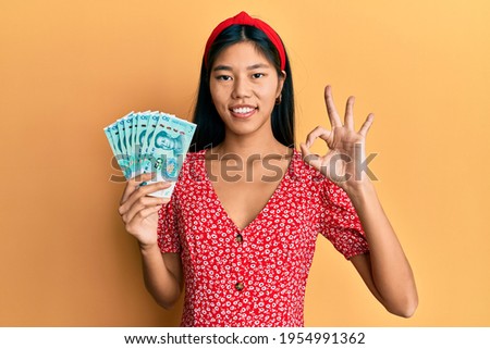 Young chinese woman holding 50 yuan chinese banknotes doing ok sign with fingers, smiling friendly gesturing excellent symbol 