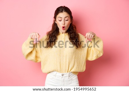 Excited cute girl checking out promo offer on bottom, pointing fingers down and saying wow, being impressed with advertisement, standing over pink background