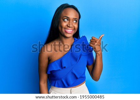 Young african woman wearing casual clothes over blue background smiling with happy face looking and pointing to the side with thumb up. 
