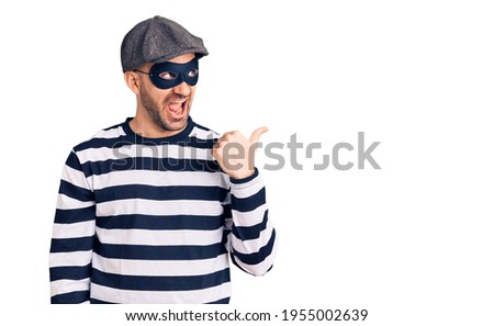 Young handsome man wearing burglar mask smiling with happy face looking and pointing to the side with thumb up.  Royalty-Free Stock Photo #1955002639