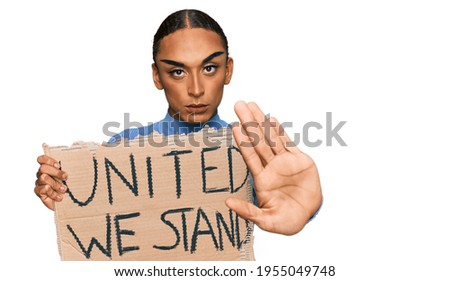 Hispanic transgender man wearing make up and long hair holding united we stand banner with open hand doing stop sign with serious and confident expression, defense gesture 
