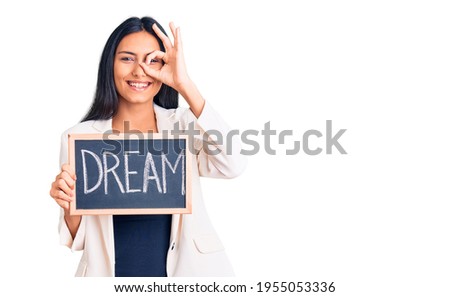 Young beautiful latin girl holding blackboard with dream word smiling happy doing ok sign with hand on eye looking through fingers 
