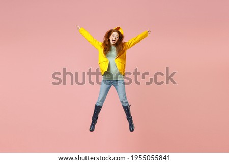 Full length redhead excited young woman in yellow waterproof raincoat outerwear jump high outstretched hands isolated on pastel pink background studio Outdoor lifestyle wet fall weather season concept