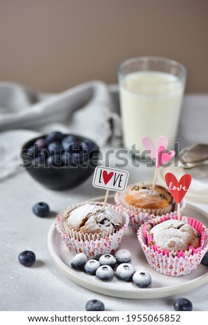 Blueberry muffins in paper tins, sprinkled with powdered sugar, with I love you flags, on a white marble board, in the background a bowl with blueberries and a glass of milk.