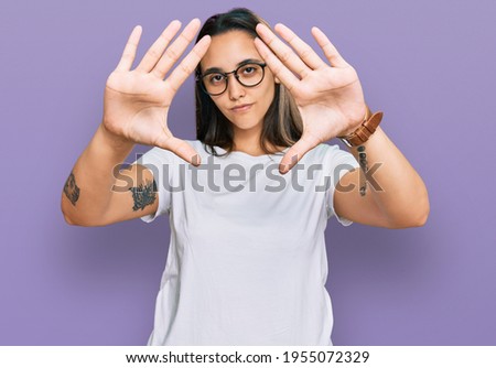 Young hispanic woman wearing casual white t shirt doing frame using hands palms and fingers, camera perspective 
