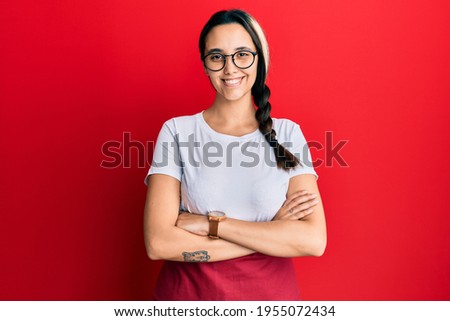 Young hispanic woman wearing professional waitress apron happy face smiling with crossed arms looking at the camera. positive person. 