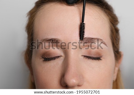 Beautician brushing woman's eyebrows before tinting on light grey background, closeup