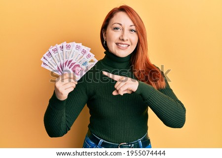 Beautiful redhead woman holding 50 mexican pesos banknotes smiling happy pointing with hand and finger 