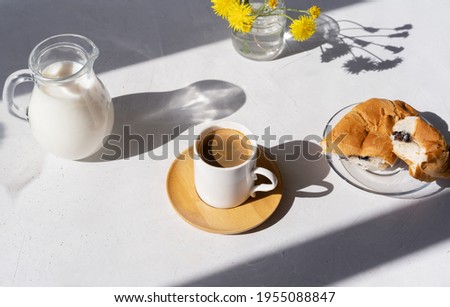 Cup of black fresh coffee with croissant, jar of milk and dandelions on the light background with shadows