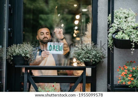 Business concept, caucasian man holding open sign indoors in shop. Young man flower center employee in apron standing in garden center. Millennial guy in studio with plants in pots turns nameplate