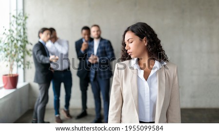 Sexism. Group Of Businessmen Whispering Behind Back Of Victimized Latin Businesswoman In Modern Office. Females Discrimination Problem, Workplace Bullying Concept. Panorama, Selective Focus Royalty-Free Stock Photo #1955099584