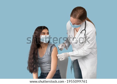 Medical doctor or nurse giving coronavirus vaccine shot to Indian teen girl over blue studio background. Female teenager in face mask getting covid-19 vaccine, participating in immunization campaign Royalty-Free Stock Photo #1955099761
