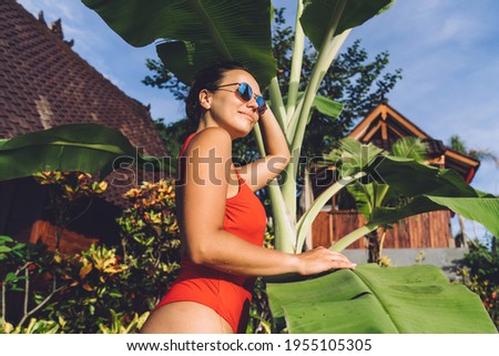 Low angle side view of glad young female in swimwear and sunglasses standing near exotic palm and looking away near bungalows on resort