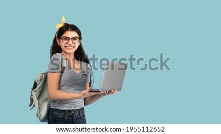 Portrait of smiling Indian teen girl using laptop computer, studying online on blue studio background, banner design with copy space. Happy teenager lady participating in educational webinar Royalty-Free Stock Photo #1955112652