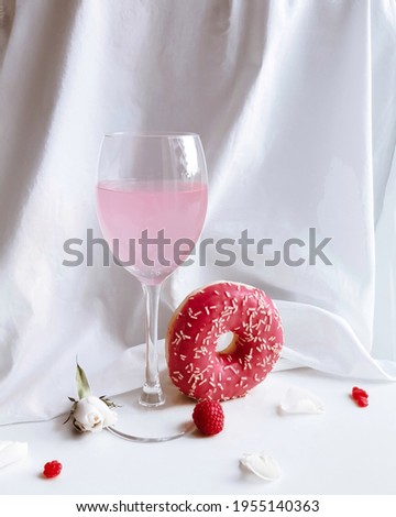 A glass of pink lemonade surrounded by a donut, raspberries, rose petals and a rosebud on a white background.