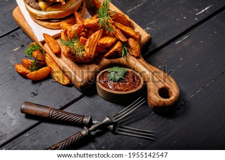 Appetizing fried potato wedges and burger on a wooden cutting board. Big portion of baked potatoes with herbs and tomato sauce. Fast food foodporn photography. Royalty-Free Stock Photo #1955142547
