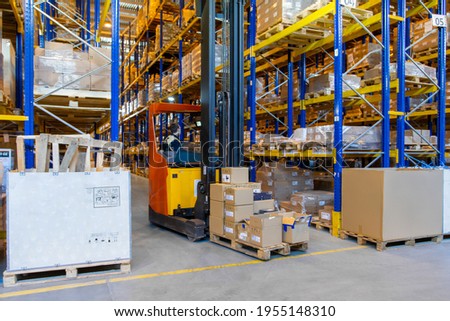 Warehouse worker driving a forklift truck full of goods between high blue yellow steel shelves inside warehouse