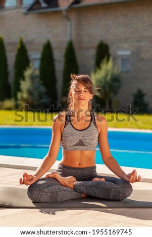 Beautiful active young woman doing yoga lotus position, practising yoga outdoors by the swimming pool on a sunny summer day