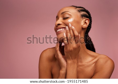 Beautiful woman eyes closed while applying moisturizer to her face. Woman applying face cream against pink background. Royalty-Free Stock Photo #1955171485