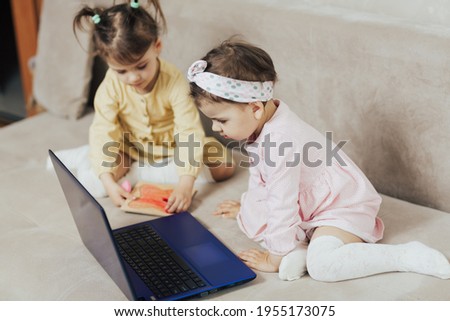 Children spending a time together at home in quarantine. One little girl drawing on wooden board, second girl watching cartoons on the laptop.