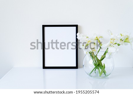 Blank black frame mockup. Fresh white irises flowers in sphere shaped glass vase on white table. White background, minimal and elegant space