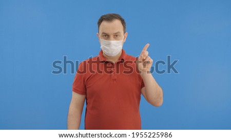 Young man wearing a medical mask protecting from the epidemic. With his finger, he points to his mask, then the ad space above it.Indoor studio shot isolated on blue background. 