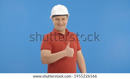 Young handsome civil engineer wearing a white hard hat. Engineer wears hard hat on isolated blue background.Thumbs up sign. Indoor studio shot isolated on blue background. 