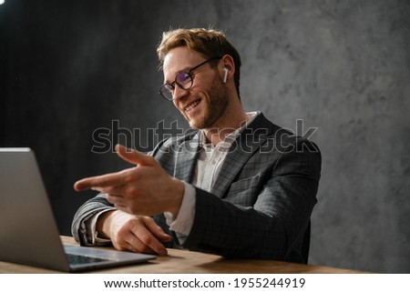 The smiling man in a jacket and glasses talking by video call on a laptop gesturing with his hands while sitting at the table in the office