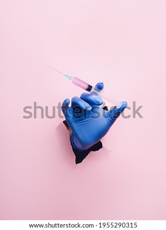 Doctor hand with surgical gloves breaking through the wall and holding syringe on pink background. Creative mass vaccination concept. Covid-19, influenza or flu global pandemic immunization. Royalty-Free Stock Photo #1955290315