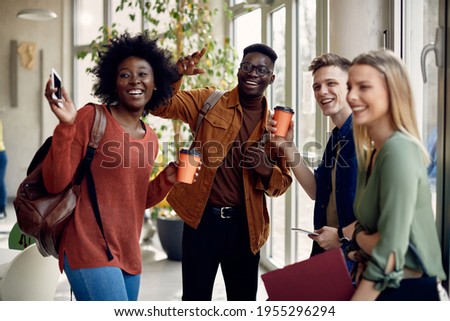 Cheerful group of college friends having fun and waving to someone while standing in a hallway. Focus is on male African American student.  Royalty-Free Stock Photo #1955296294