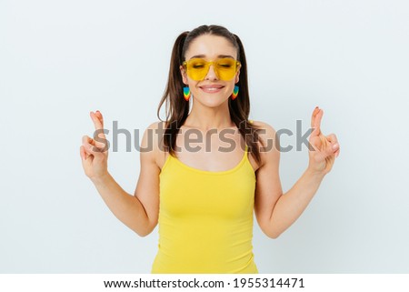 Beautiful young woman wearing summer yellow sunglasses makes wish with her eyes closed and fingers crossed while standing against white studio background.