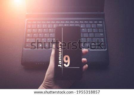 January 9th. Day 9 of month, Calendar date. Hand Holding Mobile Phone on Laptop Computer dark background with sunshine.  Winter month, day of the year concept