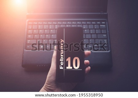 February 10th. Day 10 of month, Calendar date. Hand Holding Mobile Phone on Laptop Computer dark background with sunshine.  Winter month, day of the year concept