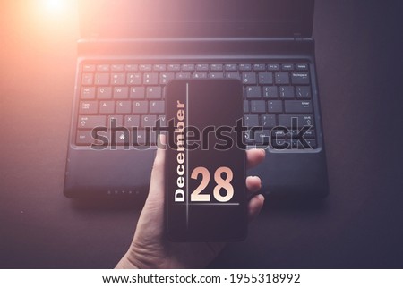 December 28th. Day 28 of month, Calendar date. Hand Holding Mobile Phone on Laptop Computer dark background with sunshine.  Winter month, day of the year concept