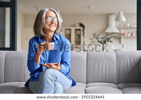 Smiling mature elder 60s woman sitting relaxing with cup of tea, coffee. Senior mid age stylish look woman with eyeglasses portrait with cup looking away at modern home. Royalty-Free Stock Photo #1955324641