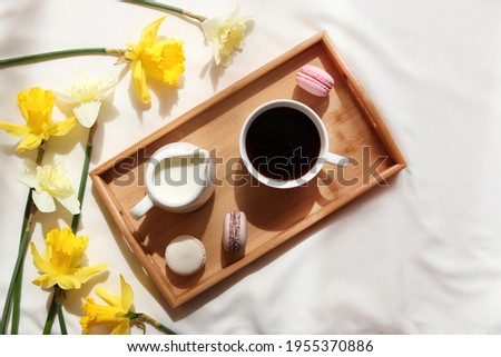 Cup of coffee with milk and French macaroons on a wooden tray and yellow daffodil flowers. Cozy home interior. Spring concept. Breakfast in bed.