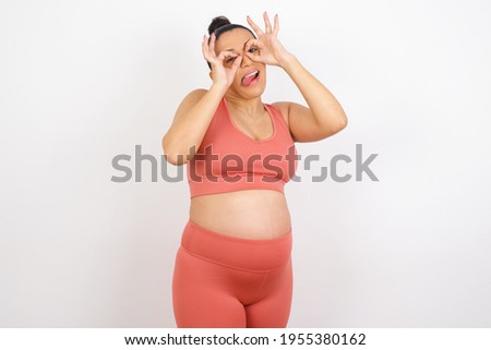 Beautiful pregnant woman in sports clothes against white background doing ok gesture like binoculars sticking tongue out, eyes looking through fingers. Crazy expression.