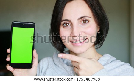 European woman with short hair smiling and showing a mobile phone with green screen and her index finger is pointing towards the phone