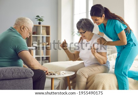 Old retired man and woman enjoy intellectual table games together. Supportive caretaker watching couple of happy senior patients play checkers. Therapy, joy, fun leisure activities in retirement home Royalty-Free Stock Photo #1955490175