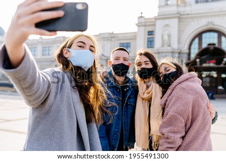 Happy millennials friends taking a selfie and wearing protective face masks. Concept of health care and the new normality. Focus on the woman on the left.