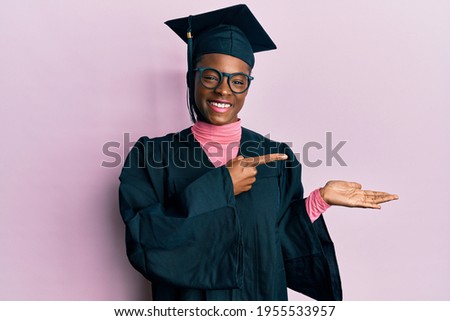 Young african american girl wearing graduation cap and ceremony robe amazed and smiling to the camera while presenting with hand and pointing with finger. 