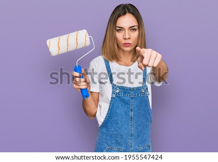 Young blonde girl holding roller painter pointing with finger to the camera and to you, confident gesture looking serious 