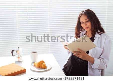 Side view of happy pregnant woman reading book while having morning breakfast with coffee and croissants on background of blinds. Good morning concept and pleasant lunch break
