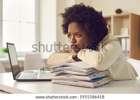 Bored unsatisfied young woman waiting for working day to end. Tired employee dissatisfied she has to work extra hours and do all paperwork sitting at office desk pouting lips with sad face expression Royalty-Free Stock Photo #1955586418