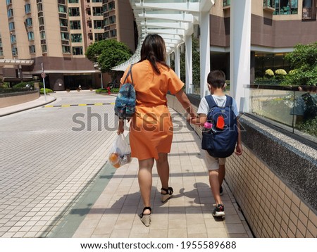 Mother or domestic helper picks up son or kid of primary school student, carrying school bag return home in afternoon, walking along street in Hong Kong on sunny day Royalty-Free Stock Photo #1955589688