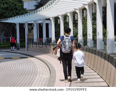 Mother or domestic helper hold hand of son or kid of kindergarten or primary school student, carrying school bag walk to or from school in  street in Hong Kong on sunny day Royalty-Free Stock Photo #1955590993