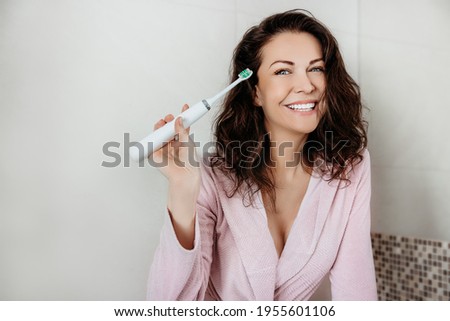 Pretty woman smiling and holding sonic toothbrush dressed in bathrobe