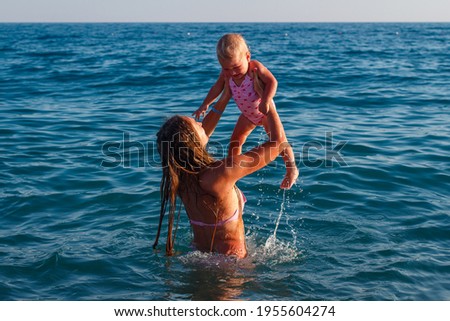 Young woman dips a one-year-old baby girl in a pink swimsuit into the sea