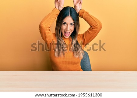 Young hispanic woman wearing casual clothes sitting on the table posing funny and crazy with fingers on head as bunny ears, smiling cheerful 