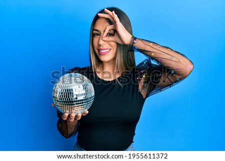 Young hispanic woman holding shiny disco ball smiling happy doing ok sign with hand on eye looking through fingers 